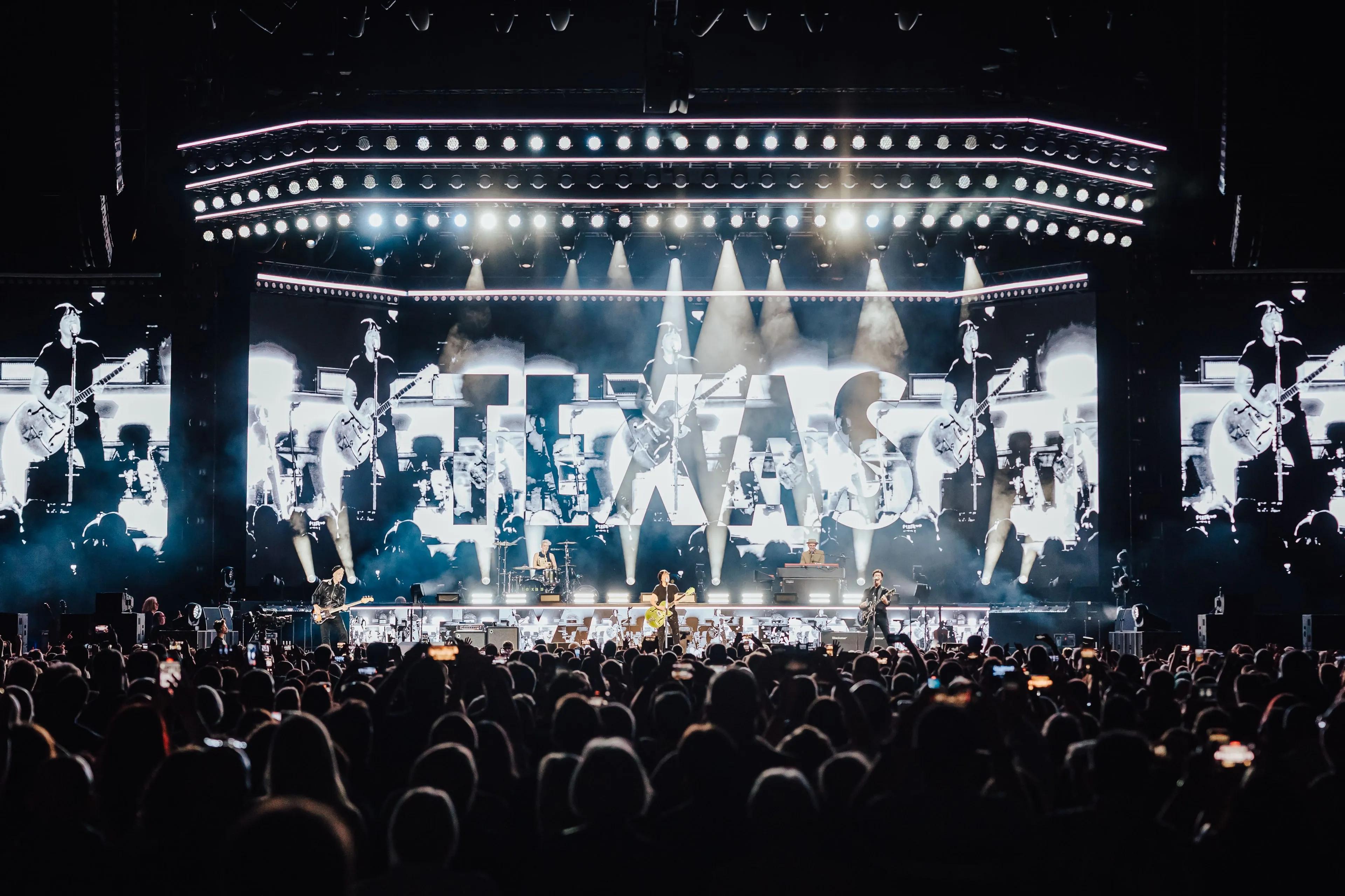 TEXAS on stage in front of a wide LED wall showing the band logo over black-and-white live camera footage, with a lighting rig above